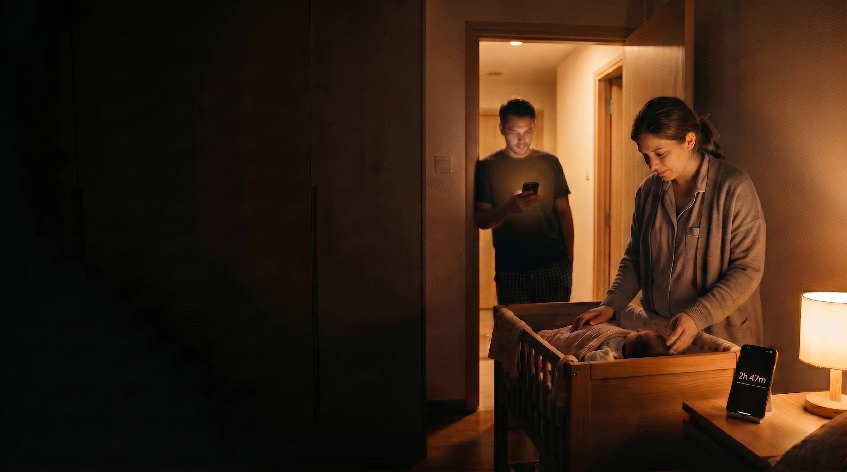 A mother settles the baby in the bassinet while her partner checks the same countdown on his phone in the doorway